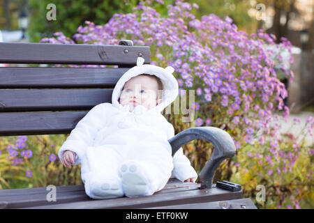 Little Baby girl in un bianco doposci seduta su una panchina accanto a un fiore violaceo bush Foto Stock