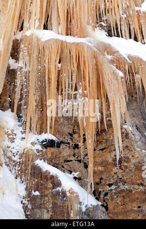 Lunga appuntita ghiaccioli essere appeso ad una parete di roccia sono colorate di arancione da ossido di ferro nel suolo. Foto Stock