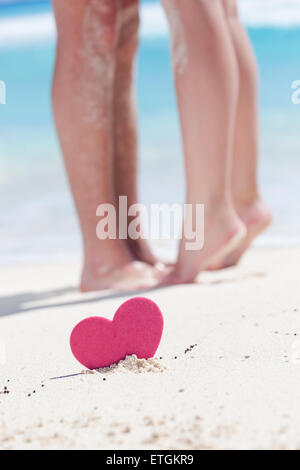 A piedi nudi gambe femmina in piedi in punta di piedi uomo di morchie sulla spiaggia con mare turchese sfondo, decorate cuore rosa oggetto. Rom Foto Stock
