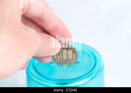 Una mano d'uomo mettendo a 500 Yen giapponese moneta in una banca blu con uno sfondo bianco. Foto Stock