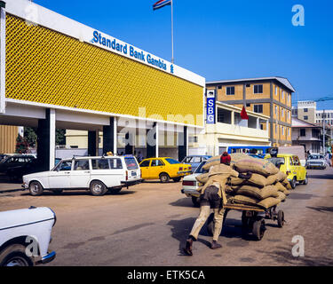 La Standard Bank Gambia edificio, uomo spingendo il carrello consegna caricate con sacchetti, Banjul (Gambia, Africa occidentale Foto Stock