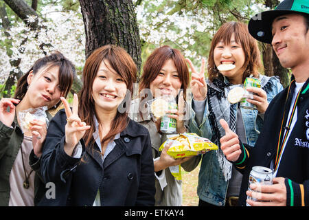 Giappone, Shukugawa. Primavera fiore ciliegio partito. Quattro giovani donne e un uomo che si affaccia spettatore, con bevande e cibo in mani e dando un gesto di pace. Foto Stock