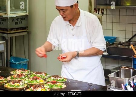 Chef giapponese, preparazione okonomiyaki su un piatto caldo nella cucina di un ristorante. I pancake saporiti che cucinano mentre cucinano aggiungono i condimenti. Kyoto. Foto Stock