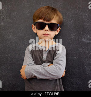 Ritratto di Little Boy indossando occhiali da sole guardando la fotocamera con le braccia incrociate. Piccolo bambino di fronte a una lavagna in posa per Foto Stock