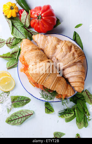 Croissant sandwich con salmone salato sulla piastra bianca, servito con insalata fresca foglie di limone, il sale di mare e le verdure sopra la luce Foto Stock