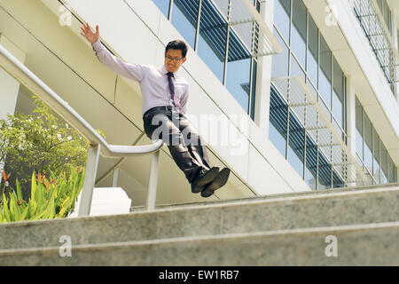 Giovani e happy businessman cinesi al piano inferiore scorrevole su binari di Office per le scale. L'uomo si gode la sua nuova promozione al lavoro Foto Stock