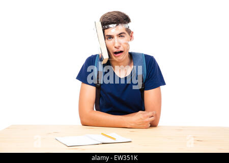 Studente di sesso maschile con libro legato alla sua testa seduto al tavolo isolato su uno sfondo bianco Foto Stock