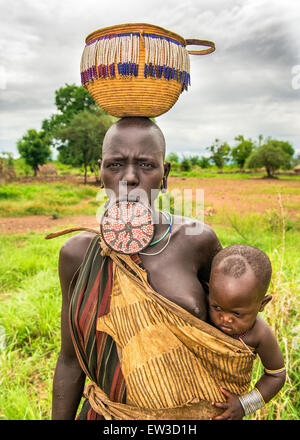 Una donna dalla tribù africana Mursi con labbro grande piastra che porta il suo bambino. Foto Stock