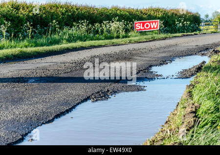 Riempito di acqua buca sulla strada di un paese con un 'Slow' firmare in background Foto Stock