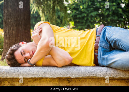 Attraente giovane uomo dorme sul banco in pietra all'aperto nel parco della città durante il giorno Foto Stock
