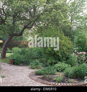 Sentiero di ghiaia accanto al mattone refilato circolare letto di erbe nel paese giardino con alberi e arbusti Foto Stock