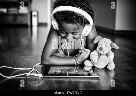 Ragazza distesa sul pavimento con il suo orsacchiotto di peluche che indossa le cuffie e guardando il suo tablet Foto Stock