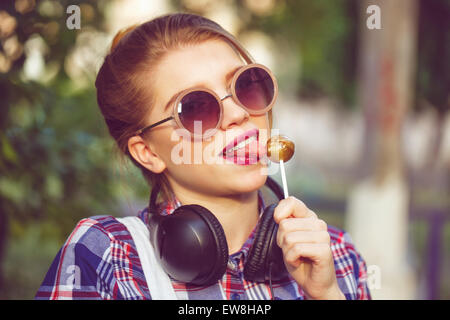 Giovane ragazza hipster ascoltando musica sulle cuffie in un parco d'estate. Close-up verticale. Ragazza leccare lecca-lecca. Tonificazione caldo. Foto Stock