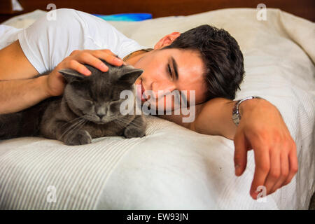 Bel giovane uomo Animal-Lover su un letto, avvolgente e coccole il suo grigio gatto domestico Pet. Foto Stock