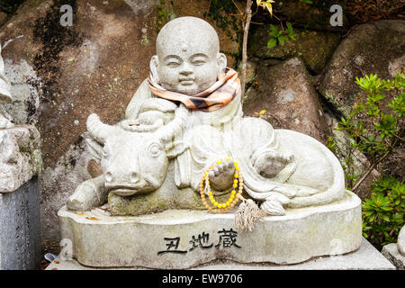 Giappone, Miyajima. Daisho-nel tempio. Piccola pietra Jizo statua della seduta monaco buddista con cappello giallo, lato seduto sulla sella sitting bull, Buffalo. Foto Stock