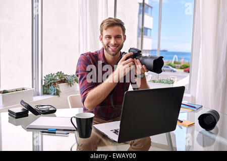 Giovane fotografo sorridente alla fotocamera con una telecamera in mani, seduto alla sua scrivania dove lavora da casa Foto Stock