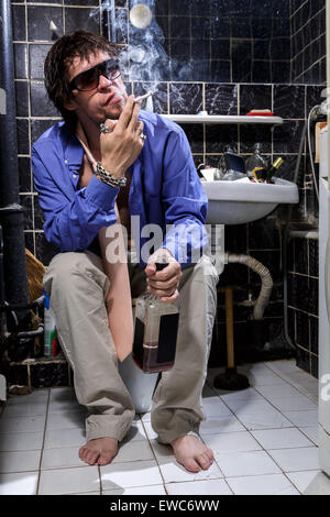 Uomo ubriaco si siede in una toilette con una bottiglia di whiskey e fumatori, immagine a colori Foto Stock