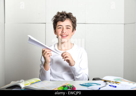 Un liscio caucasico-pelato ragazzo sta giocando con un aeroplano di carta seduto di fronte a compiti. Egli è tenendolo con la mano destra mentre la mano sinistra è la sagomatura della punta sul background industriale Foto Stock