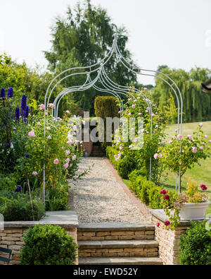Sentiero di ghiaia con letti in ferro battuto con pergola e arbusti di rosa in fiore nel giardino di Cotswold famhouse Foto Stock