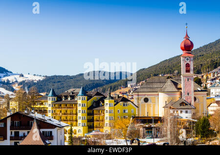 Panorama di Ortisei, villaggio italiano nelle alpi dolomitiche in Italia: case, edifici e la chiesa parrocchiale con campanile tra cime innevate e conifere Foto Stock