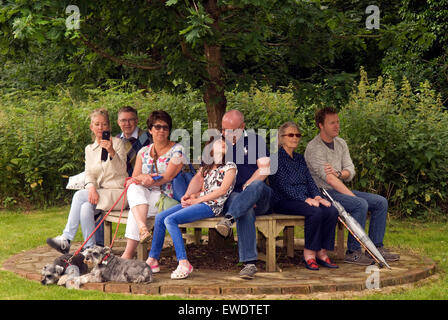 Gruppo di persone (& 2 cani) seduta su una panchina sotto un albero a guardare un estate locale comunità parade, Frensham, Surrey, Regno Unito. Foto Stock