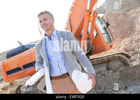 Architetto sorridente che guarda lontano tenendo blueprint e hardhat al sito in costruzione Foto Stock