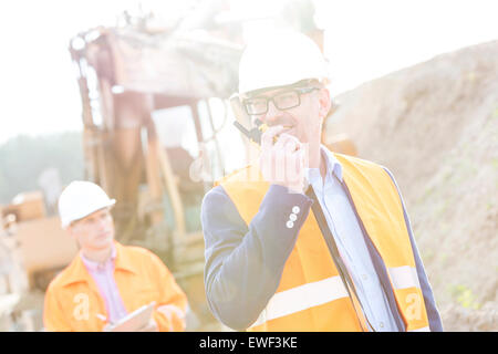 Ingegnere felice utilizzando un walkie-talkie sul sito di costruzione con il collega in background Foto Stock