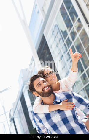 Donna felice che mostra qualcosa da uomo mentre godendo piggyback Ride in città Foto Stock