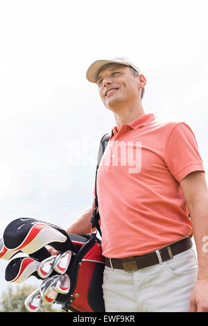 Felice uomo di mezza età che guarda lontano mentre trasportano borsa da golf contro il cielo chiaro Foto Stock