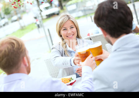 Felice imprenditrice tostare il bicchiere da birra con colleghi di sesso maschile a sidewalk cafe Foto Stock