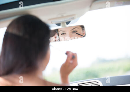 La riflessione della donna che utilizza il telefono cellulare mentre si applica il mascara in specchietto retrovisore della macchina Foto Stock