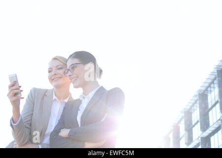 Basso angolo vista di imprenditrici utilizzando smart phone contro il cielo chiaro sulla giornata di sole Foto Stock