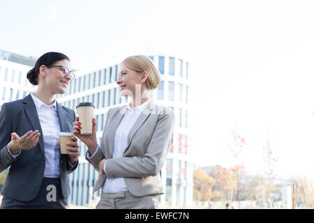 Felice imprenditrici a conversare mentre si tiene tazze monouso al di fuori di edificio per uffici Foto Stock