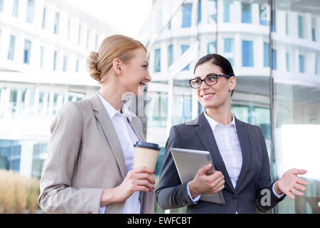 Felice imprenditrici con il tablet PC e la tazza monouso conversando al di fuori di edificio per uffici Foto Stock