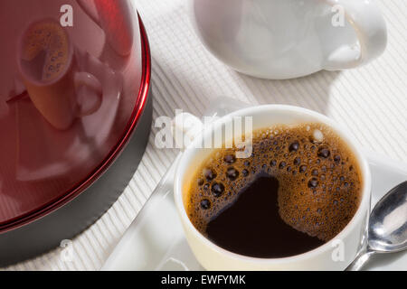 Una tazza calda di caffè nero con una riflessione sul lato di un bollitore elettrico Foto Stock