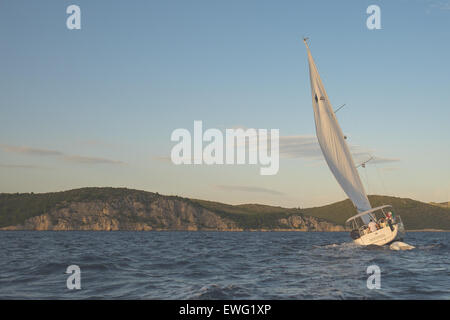 Una barca a vela naviga attraverso acque instabili con un cielo nuvoloso sopra e montagne in lontananza. La barca taglia le onde scoscese, catturando l'essenza della navigazione all'aperto in condizioni difficili. Foto Stock