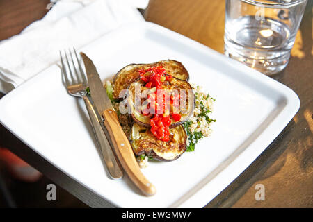 Melanzane Arrosto con cuscus su una piastra bianca di coltello e bicchiere di acqua tovagliolo bianco pomodori sul top torrefatto Foto Stock