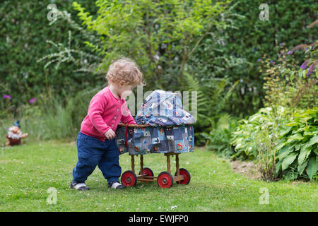 Funny curly bambina gioca con una bambola vintage passeggino in giardino Foto Stock