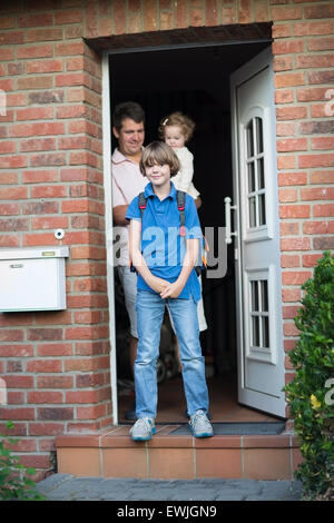 Carino ragazzo di uscire di casa per il suo primo giorno di rientro a scuola, il padre e la sorella del bambino in piedi presso la porta di casa in background Foto Stock