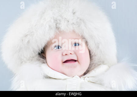 Funny Sweet Baby girl indossa un grande cappello di pelliccia e un bianco inverno tuta da neve su fondo azzurro Foto Stock