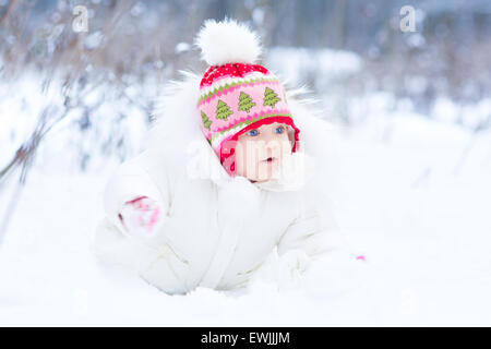 Carino divertente bambina indossa una luce bianca calda giacca invernale e un Red Hat seduto sotto un albero di natale nella neve Foto Stock