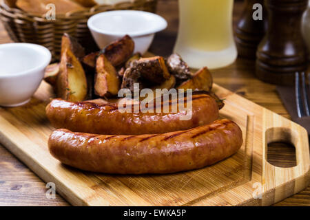 Salsicce alla griglia con patate arrosto con spezie su tavola di legno con birra chiara sul tavolo Foto Stock