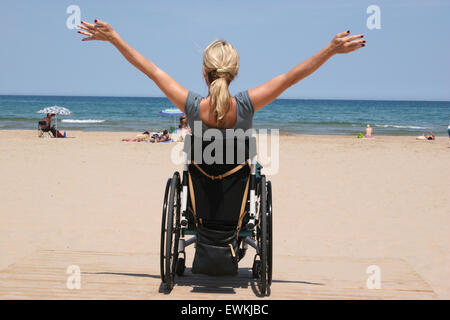 Una donna su una sedia a rotelle con le braccia aperte su una spiaggia guardando l'oceano Foto Stock