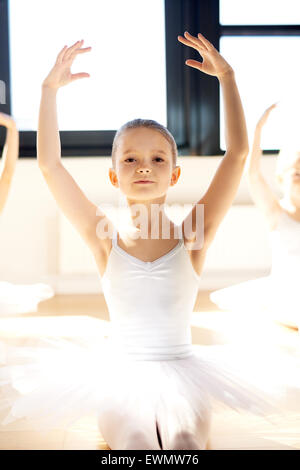 Grazioso pretty little ballerina in posa con i bracci sollevati nel suo bianco tutu in una illuminata dal sole studio di danza come lei riconosce t Foto Stock