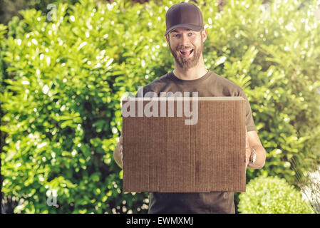 Carino barbuto ragazzo in un berretto con una felice esuberante grin facendo una consegna di una grande marrone a scatola di cartone che lo trasporta nel suo Foto Stock