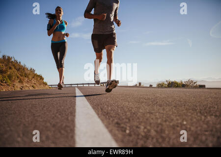 Bassa angolazione della giovane donna in esecuzione su strada con l'uomo nella parte anteriore su una mattina d'estate. Training Guide sulla strada di campagna. Foto Stock