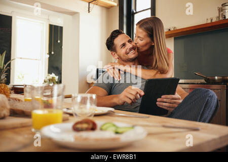 Giovane uomo in possesso di una tavoletta digitale mentre la sua ragazza abbracci di lui da dietro, dandogli un buon mattino kiss. Giovane amore giovane Foto Stock