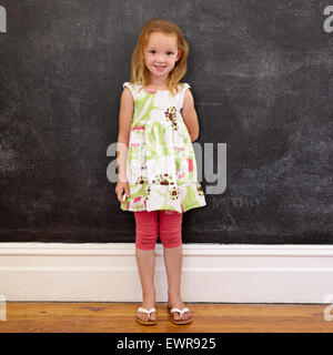 A piena lunghezza shot della bellissima bambina in piedi contro la lavagna di casa. Lei è in possesso di un chalk guardando la fotocamera. Foto Stock