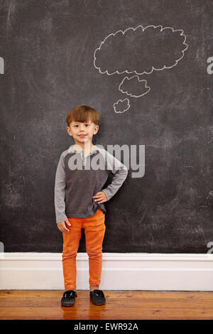 Carino piccolo ragazzo con un fumetto pensiero sulla lavagna. A piena lunghezza shot del giovane ragazzo che sta a casa con la sua mano sull'anca. Foto Stock