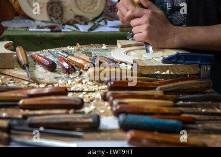 Artigiano, intagliatore di legno sta lavorando sui dettagli di un piatto di legno. Egli è in officina e sul banco di lavoro può essere visto un sacco di Foto Stock